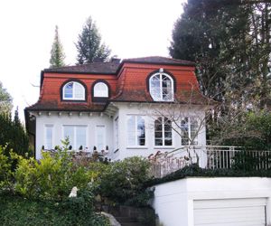 Einfamilienhaus im Grünen. Bogenfenster und Dachfenster schmücken das Haus. Mansarddach mit roten Ziegeln gedeckt. Das Landhaus hat eine Garage mit einer Terrasse darüber. Um die, vom Büro für Immobilienbewertung, bewertete Immobilie stehen grüne Bäume.