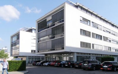 Zwei moderne Bürogebäude im Bauhausstil begutachtet vom unseren Büro für Immobilienbewertung in Frankfurt. Die weißen wie monolithe Baublocke aussehenden Immobilien haben weite Fenster. Im Erdgeschoss unter einem Überbau befinden sich gläserne Vitrinen.