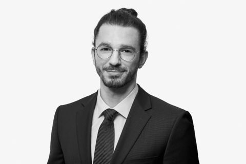 Die Fotografie des Sachverständigen für Immobilienbewertung, Bachelors of Engineering Patrick Tomiak ist in Graustufen. Der junge Mann mit Brille und zusammengebundenen Haaren ist in Businessanzug mit Krawatte gekleidet.